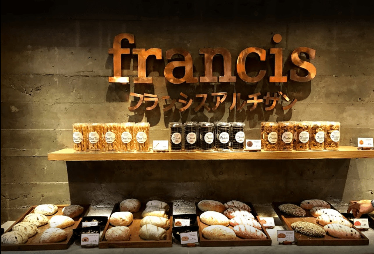  Loaf and sliced breads on wooden shelves with a variety of cookies in glass jars above them, with the words 'Francis' and 'Francis Artisan' on the wall behind.